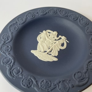 May include: A blue and white ceramic plate with a raised design of three women in a circle. The plate has a decorative border.