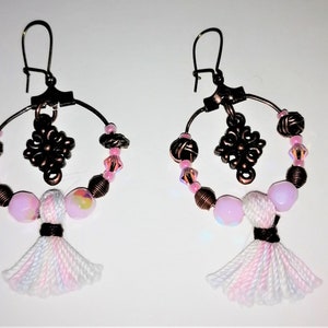Pode incluir: Um par de brincos de argola com contas rosa e brancas, um amuleto de cobre e um tassel rosa e branco.