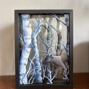 May include: A rectangular black shadow box frame containing a layered paper art scene. The display features white and blue birch trees with bare branches and a brown deer with antlers standing among the trees, creating a three-dimensional effect.