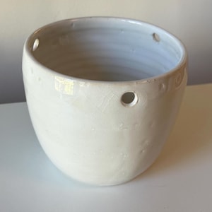 May include: A white ceramic planter with a glossy, crackled glaze finish. The round vessel features a series of small, evenly spaced circular holes near the upper rim, designed for hanging. The surface has a subtle, textured, handmade appearance.