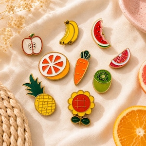 Könnte beinhalten: Eine Sammlung von Emaille-Anstecknadeln mit verschiedenen Obst- und Gemüsemotiven, darunter ein roter Apfel, eine gelbe Banane, eine Wassermelonenscheibe, eine Orangenscheibe, eine Karotte, eine Kiwischeibe, eine Ananas und eine gelbe Sonnenblume.