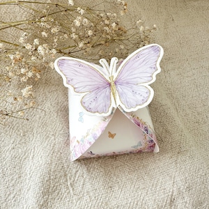 Könnte beinhalten: Eine kleine, weiße Geschenkbox mit einem lila Schmetterlingsdesign. Die Box hat ein Blumenmuster und ist mit goldenen Akzenten verziert.
