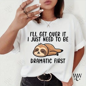 Puede incluir: Camiseta blanca con el texto "I'll get over it, I just need to be dramatic first" y una ilustración de un perezoso. El perezoso es marrón y beige, con los ojos cerrados. La camiseta tiene mangas enrolladas.