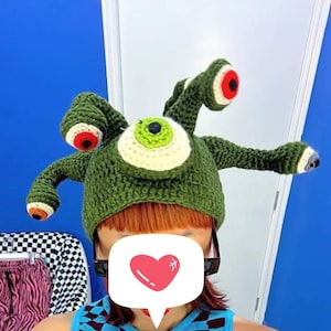 May include: A green crocheted hat shaped like a monster with three eyes. The hat has a large green eye in the centre and two smaller red eyes on the sides. The hat has long green tentacles coming out of the top.