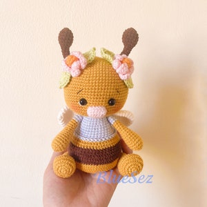 May include: A crocheted bee toy with a yellow and brown striped body, purple wings, and a pink nose. The bee is wearing a flower crown with pink and green flowers.
