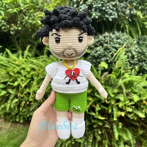 Puede incluir: Mu&ntilde;eca de crochet con pelo negro, una camisa blanca con un coraz&oacute;n rojo y una cara triste, pantalones cortos verdes y zapatos blancos.