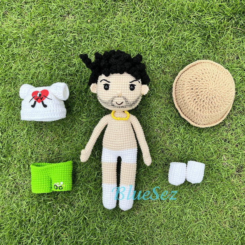 Puede incluir: Mu&ntilde;eca de crochet con cabello negro, bigote y barba. La mu&ntilde;eca lleva ropa interior blanca, calcetines blancos y un collar amarillo. La mu&ntilde;eca est&aacute; rodeada de prendas de crochet, incluyendo una camisa blanca con un coraz&oacute;n rojo, pantalones cortos verdes, un sombrero marr&oacute;n y calcetines blancos.