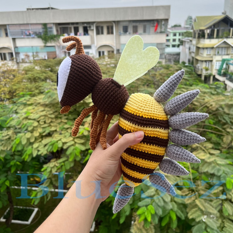 Puede incluir: Un juguete de abeja de crochet con rayas marrones y amarillas, alas grises y un ojo blanco. La abeja est&aacute; en la mano de una persona.