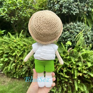 Puede incluir: Una mu&ntilde;eca de crochet con una camisa blanca, pantalones cortos verdes y un sombrero marr&oacute;n. La mu&ntilde;eca est&aacute; de pie sobre una mano.