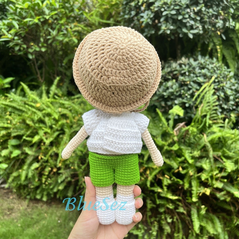 Puede incluir: Una mu&ntilde;eca de crochet con una camisa blanca, pantalones cortos verdes y un sombrero marr&oacute;n. La mu&ntilde;eca est&aacute; de pie sobre una mano.
