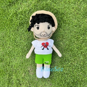 Puede incluir: Una mu&ntilde;eca de crochet que lleva un sombrero de paja, una camisa blanca con un dise&ntilde;o de coraz&oacute;n rojo y pantalones cortos verdes. La mu&ntilde;eca tiene el pelo negro y una barba marr&oacute;n.