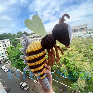 Puede incluir: Un juguete de peluche de abeja tejido a crochet con rayas marrones y amarillas, un ala verde claro en forma de coraz&oacute;n y un aguij&oacute;n gris. La abeja est&aacute; siendo sostenida por una mano.