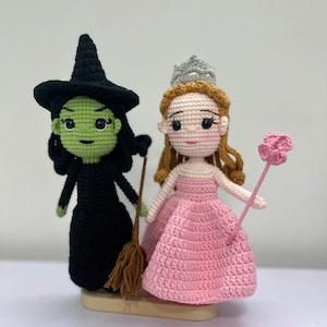 May include: Crocheted dolls of the Wicked Witch of the West and Glinda the Good Witch. The witch is in a black dress and hat, holding a broom. Glinda wears a pink dress and a silver crown, holding a pink flower wand. The dolls are handmade.