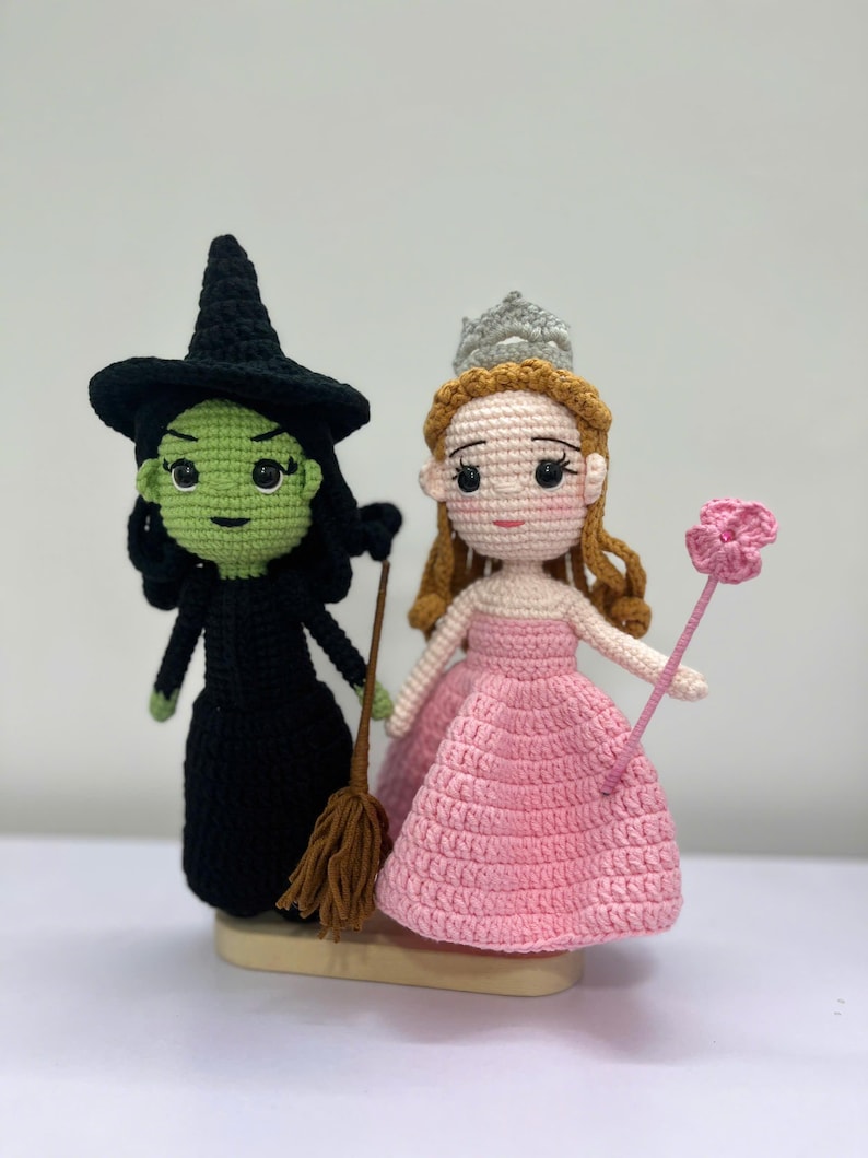 May include: Crocheted dolls of the Wicked Witch of the West and Glinda the Good Witch. The witch is in a black dress and hat, holding a broom. Glinda wears a pink dress and a silver crown, holding a pink flower wand. The dolls are handmade.
