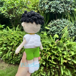 Puede incluir: Una mu&ntilde;eca de crochet con pelo negro, una camisa blanca, pantalones cortos verdes y zapatos blancos. La mu&ntilde;eca est&aacute; siendo sostenida por una mano.
