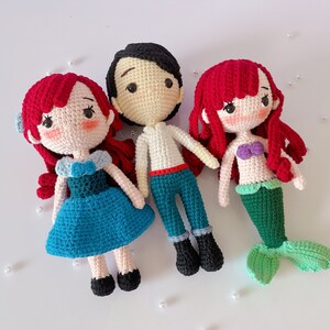 May include: Three handmade crochet dolls: two mermaids and a prince. The dolls have red hair, and the mermaid has a green tail. The prince wears blue pants and a white shirt. The dolls are made with yarn and have embroidered facial features.