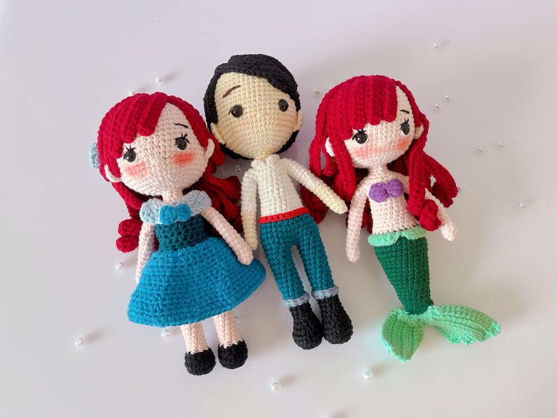 May include: Three handmade crochet dolls: two mermaids and a prince. The dolls have red hair, and the mermaid has a green tail. The prince wears blue pants and a white shirt. The dolls are made with yarn and have embroidered facial features.