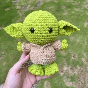 K&ouml;nnte beinhalten: Eine gr&uuml;ne Yoda-Puppe aus H&auml;kelarbeit, die eine beige H&auml;keljacke tr&auml;gt. Die Puppe hat schwarze Knopfaugen und h&auml;lt ihre H&auml;nde vor sich.