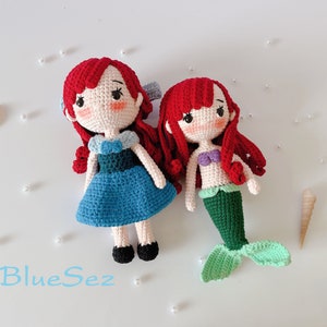 May include: Two handmade crochet dolls: one in a blue dress and the other a mermaid. Both dolls have red hair, rosy cheeks, and black eyes. The mermaid doll has a green tail and a purple top. The dolls are on a white surface with the text "BlueSez".