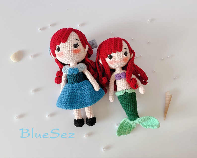 May include: Two handmade crochet dolls: one in a blue dress and the other a mermaid. Both dolls have red hair, rosy cheeks, and black eyes. The mermaid doll has a green tail and a purple top. The dolls are on a white surface with the text "BlueSez".