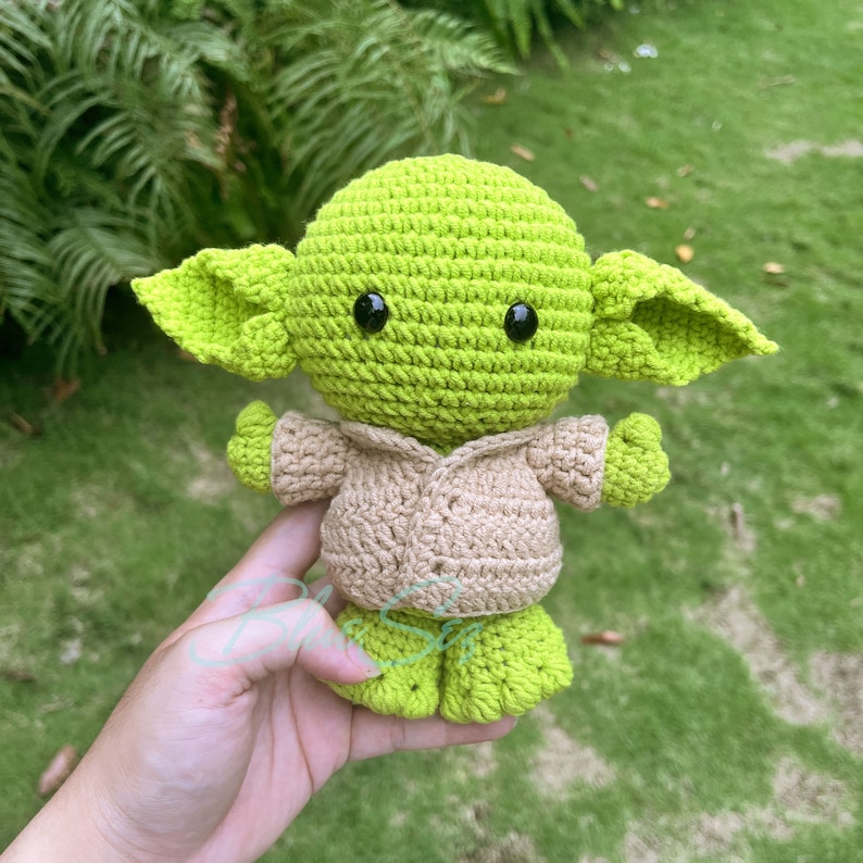 K&ouml;nnte beinhalten: Eine gr&uuml;ne geh&auml;kelte Yoda-Puppe, die ein beigefarbenes geh&auml;keltes Hemd tr&auml;gt. Die Puppe hat gro&szlig;e schwarze Augen und h&auml;lt ihre H&auml;nde vor sich.