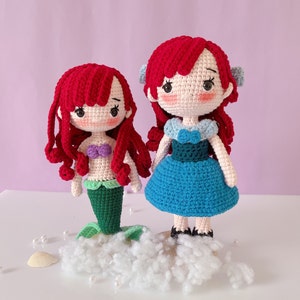 May include: Two handmade crochet dolls with red hair. One doll has a green mermaid tail and a purple top. The other doll wears a blue dress. Both dolls have light skin and are standing on a white cloud-like base.