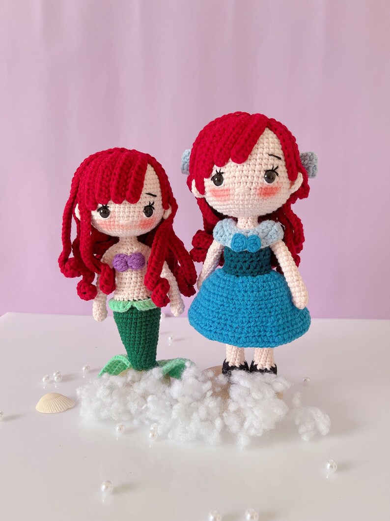 May include: Two handmade crochet dolls with red hair. One doll has a green mermaid tail and a purple top. The other doll wears a blue dress. Both dolls have light skin and are standing on a white cloud-like base.