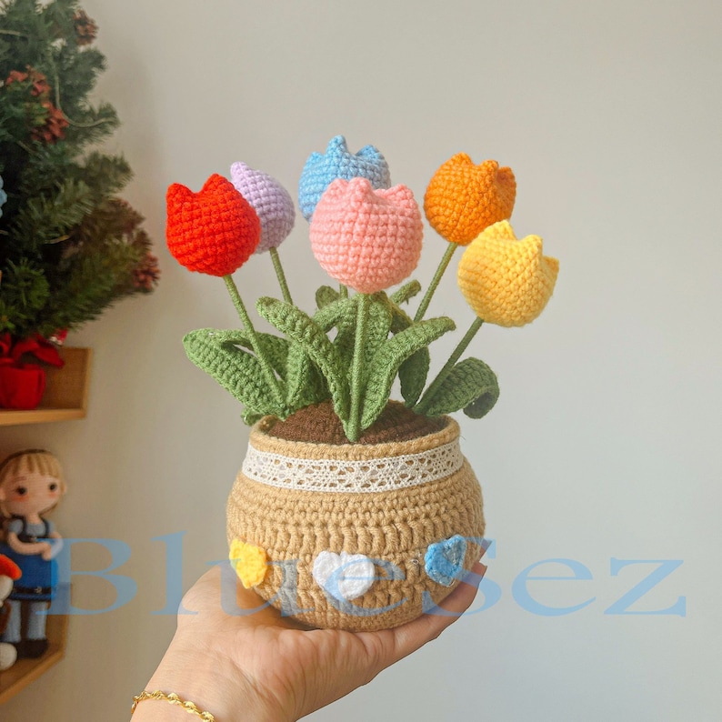 May include: A crocheted flower arrangement with six colorful tulips in a brown pot with a white lace trim. The pot is decorated with colorful heart-shaped buttons.