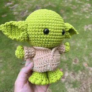 K&ouml;nnte beinhalten: Eine gr&uuml;ne geh&auml;kelte Yoda-Puppe, die eine braune geh&auml;kelte Jacke tr&auml;gt. Die Puppe hat gro&szlig;e schwarze Augen und einen kleinen Mund.