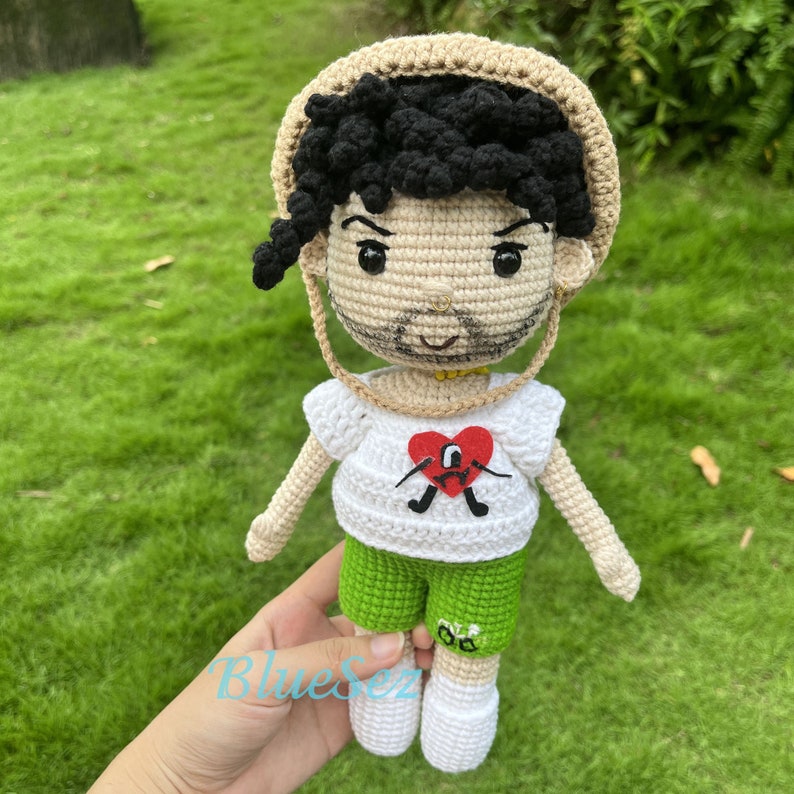 Puede incluir: Mu&ntilde;eca de crochet con cabello negro rizado, vistiendo una camisa blanca con un dise&ntilde;o de coraz&oacute;n rojo, pantalones cortos verdes y un sombrero de paja.