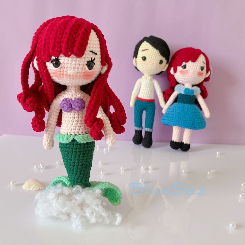 May include: Crocheted dolls of a mermaid with red hair, a purple seashell top, and green tail, and two other dolls. The mermaid doll is the largest, with a white cloud-like base. The other dolls are a male doll in blue pants and a female doll in a blue dress.