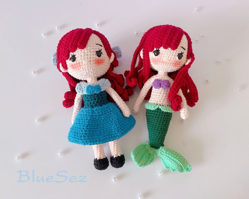 May include: Two handmade crochet dolls with red hair. One doll wears a blue dress and black shoes, the other is a mermaid with a green tail and a purple top. The dolls have rosy cheeks and are on a white surface.