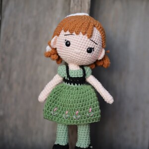 May include: A crocheted doll with brown hair, a green dress, and black shoes. The doll has a sweet expression and is standing on a red surface.