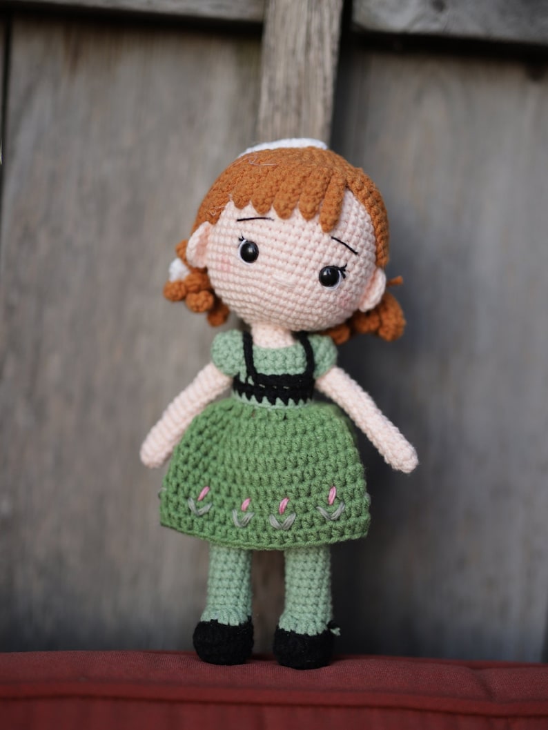 May include: A crocheted doll with brown hair, a green dress, and black shoes. The doll has a sweet expression and is standing on a red surface.