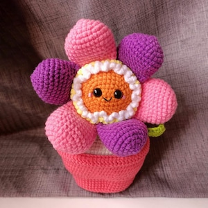 Puede incluir: Una maceta de ganchillo con una cara sonriente. La maceta es rosa, morada y naranja con un borde blanco. La maceta está sentada sobre una superficie gris.