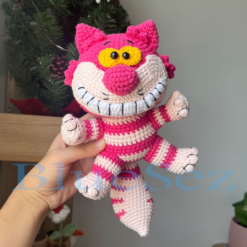 May include: A pink and white striped crocheted cat with a wide grin and yellow eyes. The cat has a long tail and is holding its paws up.