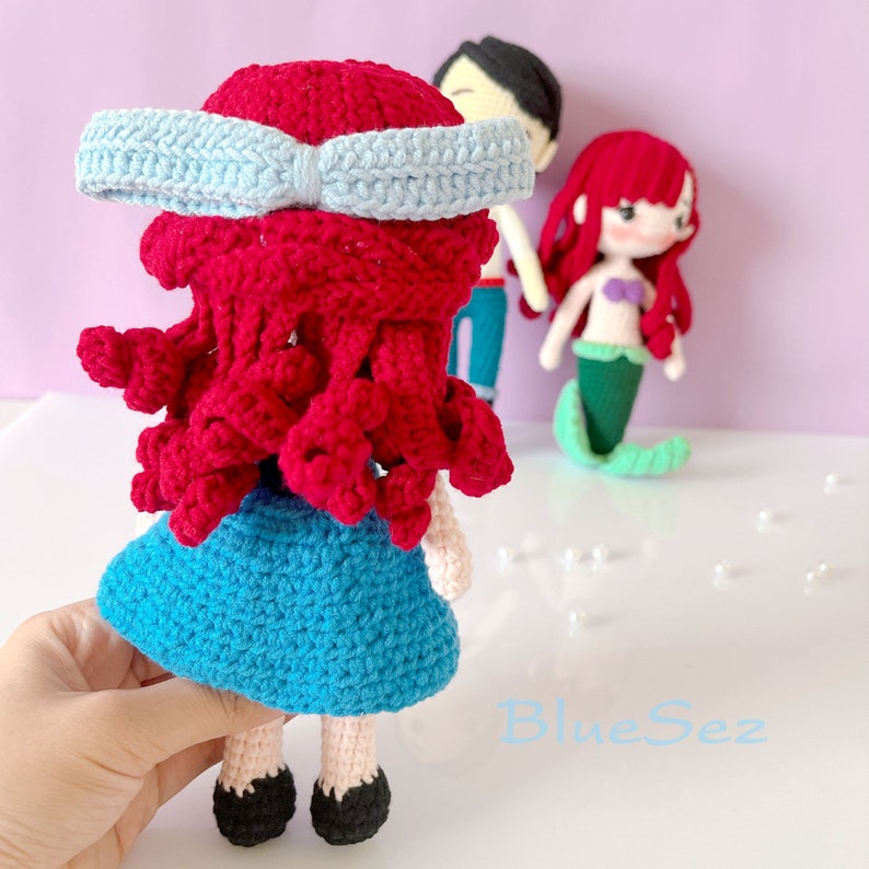 May include: Crocheted doll of a mermaid with red hair, a blue dress, and a light blue bow. Other crocheted dolls are in the background. The doll is handmade and has black shoes. The doll is a handmade item.
