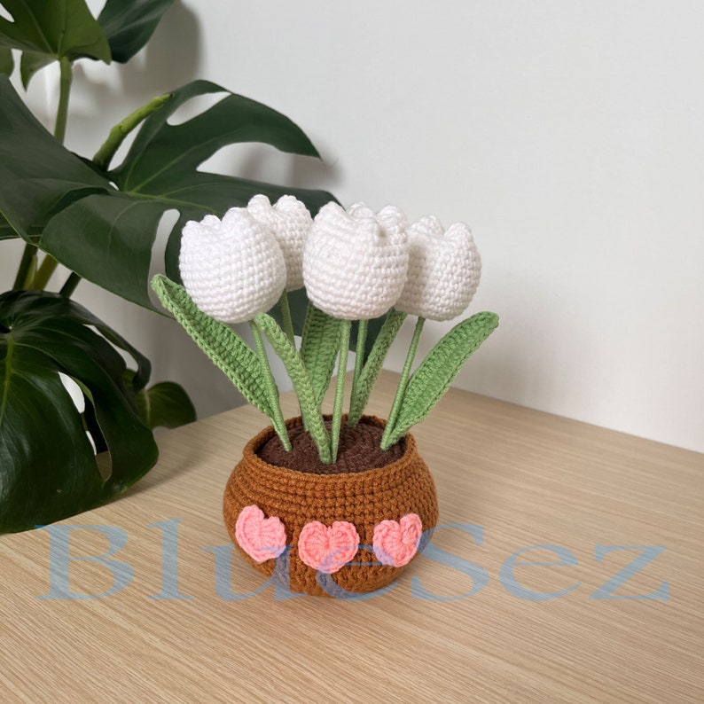 May include: A crocheted bouquet of white tulips in a brown pot with pink hearts around the base.