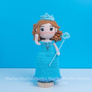 May include: Crocheted doll of a princess wearing a blue dress, crown, and holding a wand. The doll has brown hair and rosy cheeks. The background is a solid blue. The doll is approximately 8 inches tall.