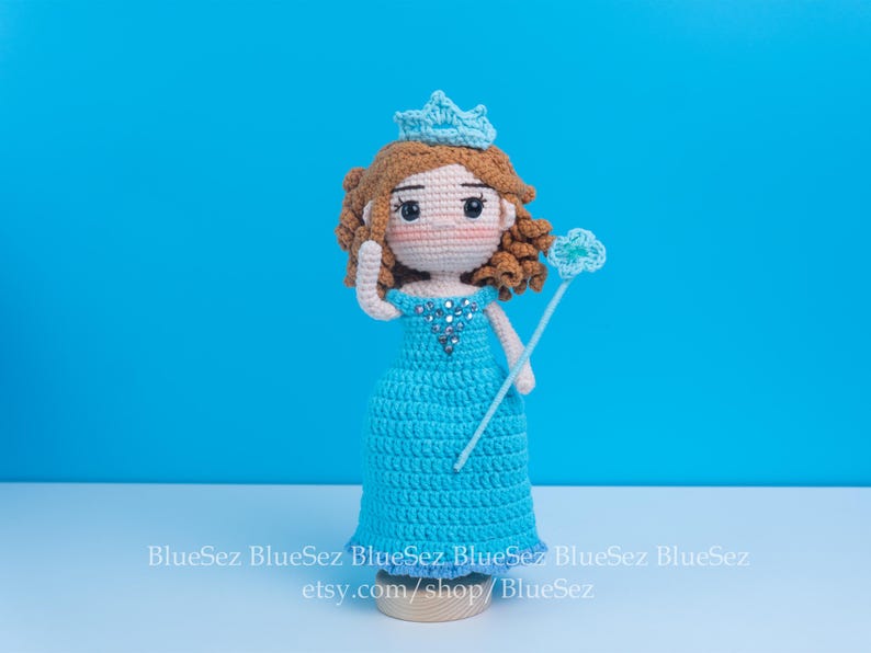May include: Crocheted doll of a princess wearing a blue dress, crown, and holding a wand. The doll has brown hair and rosy cheeks. The background is a solid blue. The doll is approximately 8 inches tall.