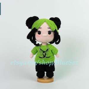 May include: Crocheted doll with a green top, black trousers, and black hair styled in space buns. The doll has a light skin tone and is standing on a wooden base. The doll's top has a black design. The text "etsy.com/shop/BlueSez" is visible.