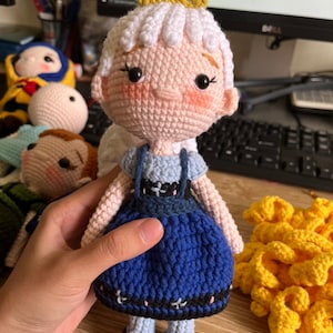 May include: A crocheted doll with a white dress, blue skirt, and a yellow crown. The doll has pink cheeks and black eyes.