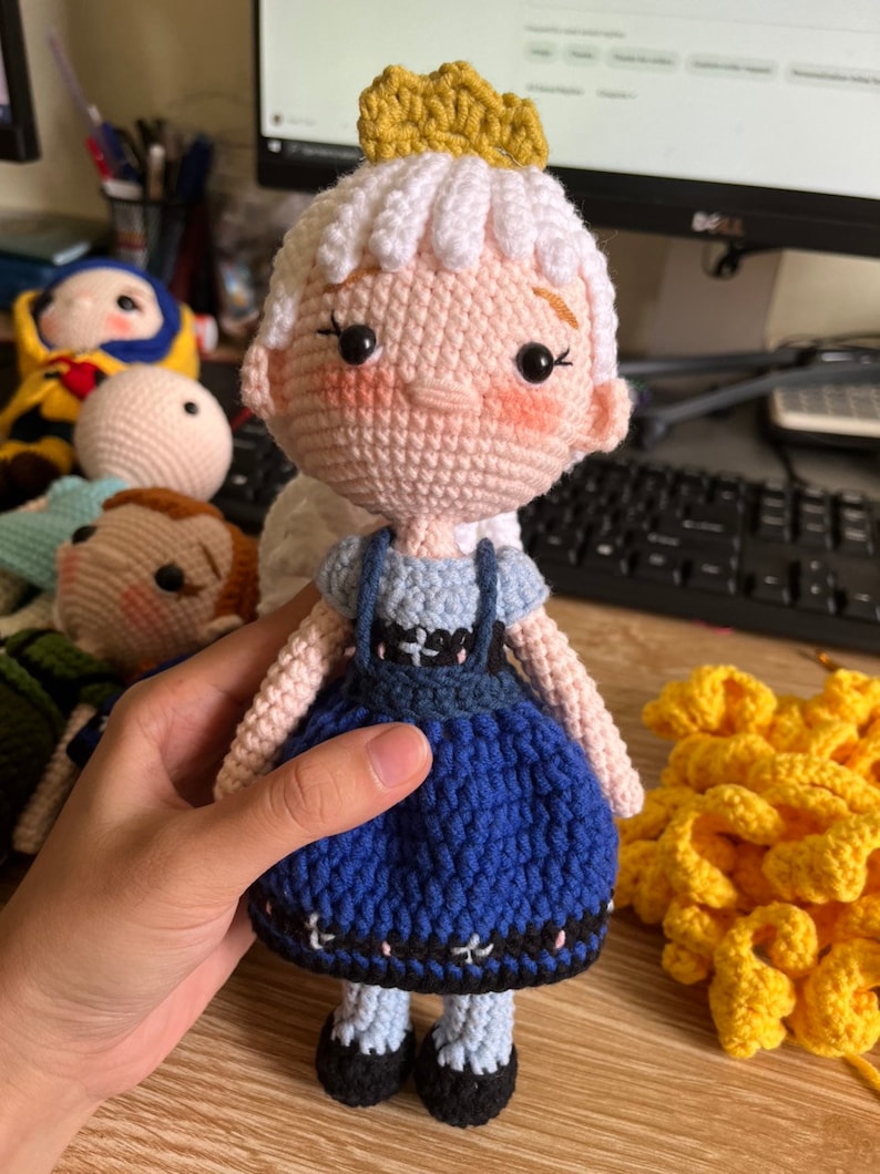 May include: A crocheted doll with a white dress, blue skirt, and a yellow crown. The doll has pink cheeks and black eyes.