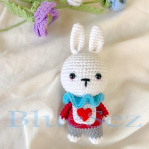 May include: White crocheted bunny rabbit wearing a red sweater with a blue collar and a red heart. The bunny has black eyes and a black nose.