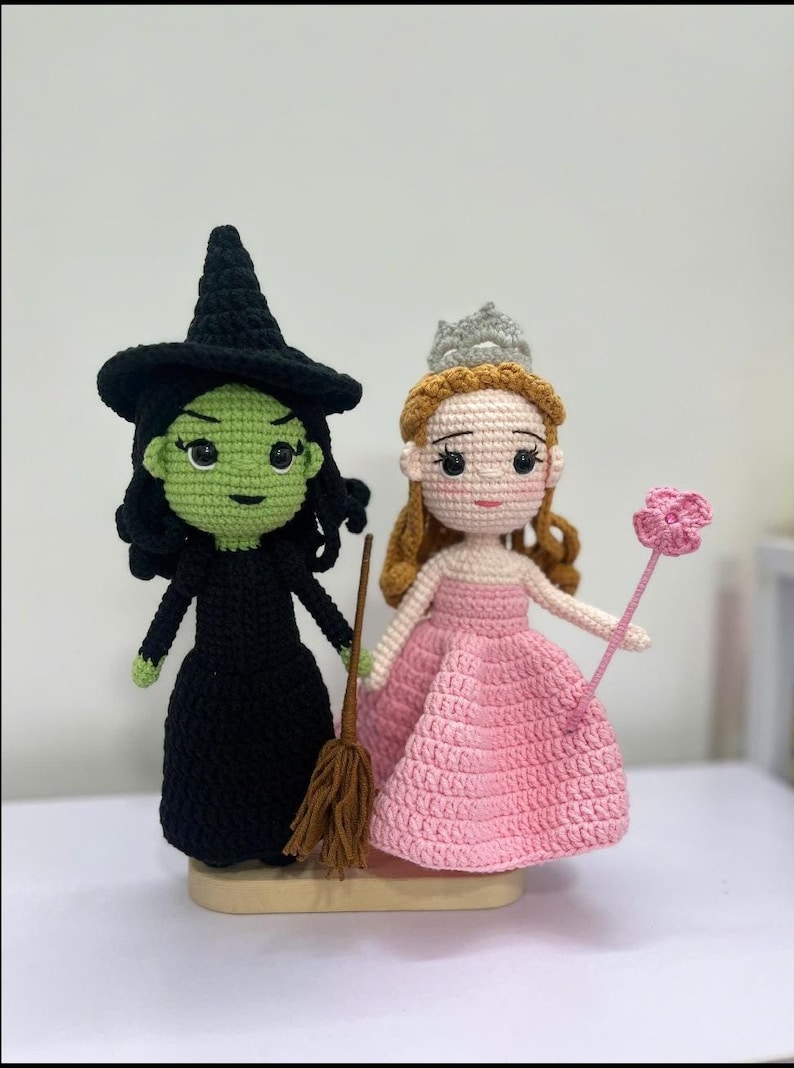 May include: Crocheted dolls of the Wicked Witch of the West and Glinda the Good Witch from The Wizard of Oz. The witch is in a black dress and hat, holding a broom. Glinda wears a pink dress and a crown, holding a pink flower wand.