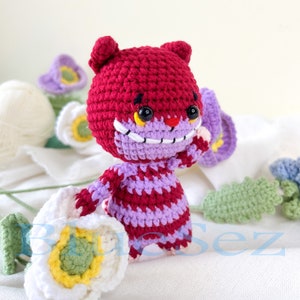 May include: A crocheted plush toy in the shape of a cat with a red and purple striped pattern. The cat has a wide grin with white teeth and yellow eyes. The cat is holding a white flower.