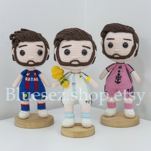 May include: Three handmade crochet dolls of soccer players. Each doll has a brown beard and hair, and is wearing a different team uniform. The dolls are standing on small wooden bases. The dolls are in a well-lit room.