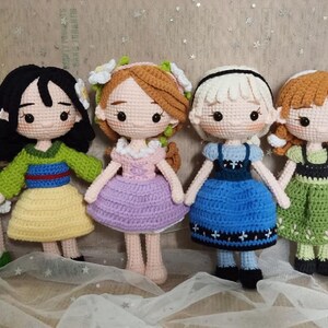 May include: Five crocheted dolls dressed as Disney princesses. The dolls are standing on a white fabric background. The dolls are wearing dresses in various colors, including green, yellow, purple, blue, and green.