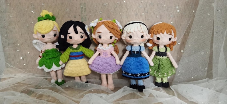 May include: Five crocheted dolls dressed as Disney princesses. The dolls are standing on a white fabric background. The dolls are wearing dresses in various colors, including green, yellow, purple, blue, and green.