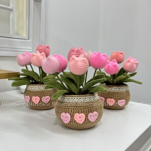 May include: Three crocheted flower arrangements in brown pots with white lace trim and pink hearts. Each pot contains pink and white crocheted tulips with green leaves.